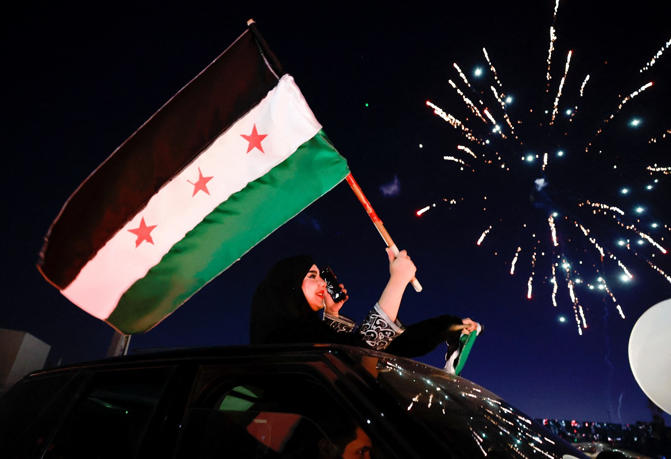 Uma mulher ergue a bandeira adotada pelos novos governantes sírios, enquanto as pessoas celebram após os combatentes do corpo governante depor Bashar al-Assad, na antiga cidade de Damasco, Síria, em 13 de dezembro de 2024. REUTERS/Ammar Awad