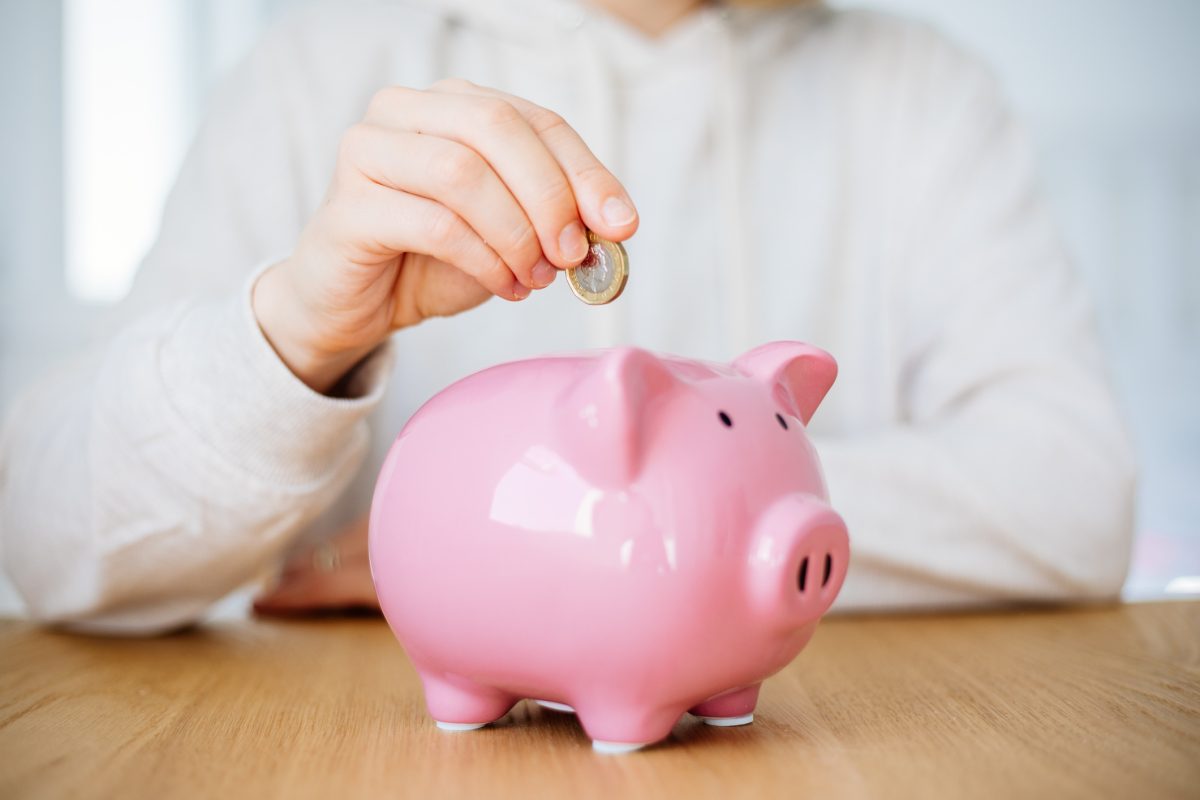 Imagem mostra mão de mulher colocando moedas em um porquinho para economizar dinheiro